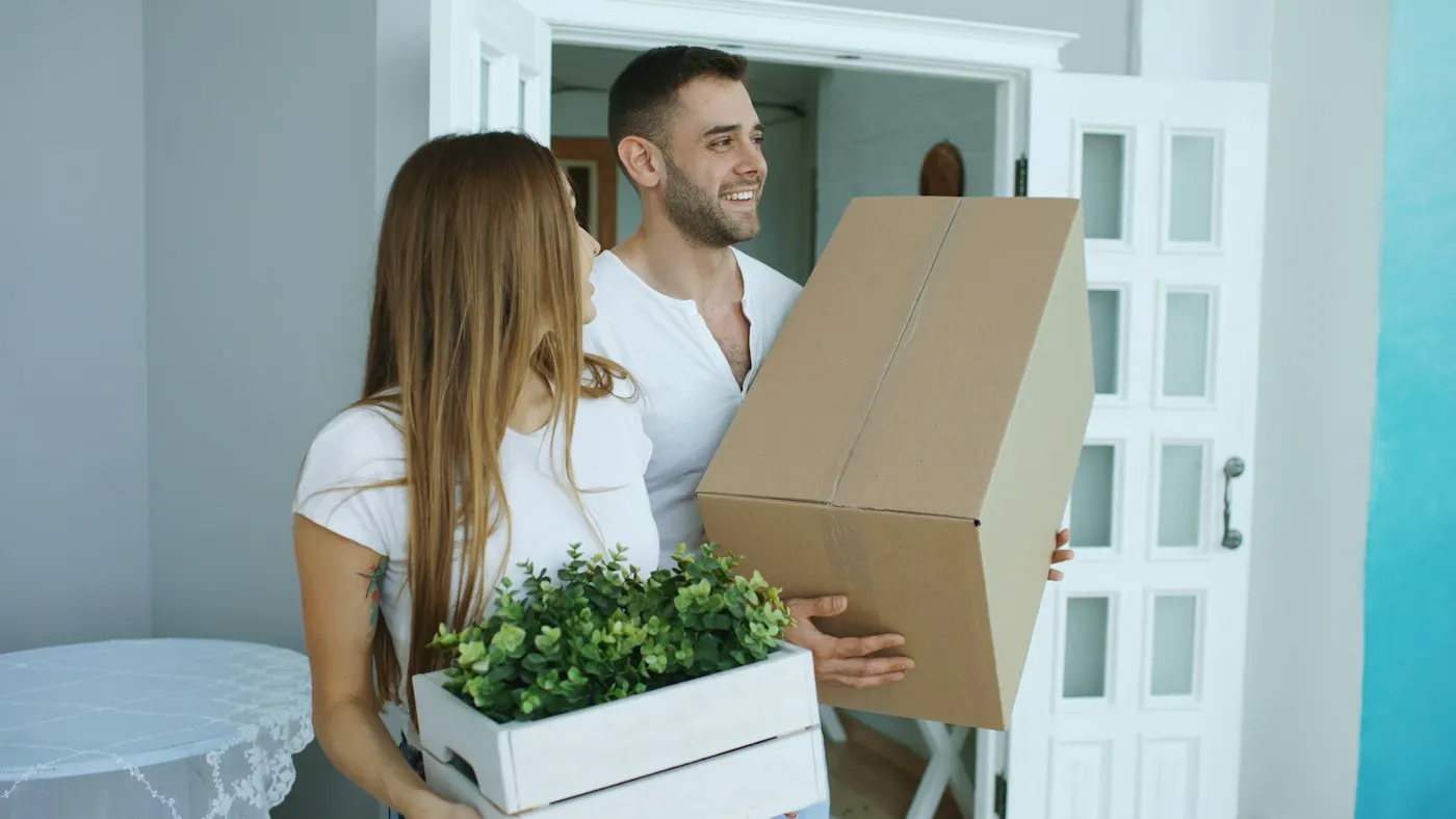 Casal sorrindo carregando uma caixa de papelão e uma planta em uma mudança